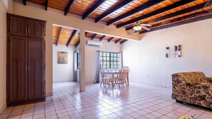 A spacious room with a tiled floor, a wooden ceiling, a dining area with chairs and a table, a sofa, and a wardrobe.