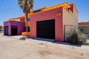 Um edifício de dois andares com porta de garagem, uma palmeira e um muro com uma placa.