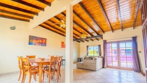 A spacious living area with a wooden ceiling, a dining table with chairs, a sofa, and a large window with curtains.