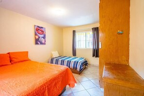 Une chambre d’hôtel avec deux lits, l’un recouvert d’une couvre-lit orange et l’autre d’une couvre-lit à rayures. Il y a une fenêtre avec des rideaux, un tableau au mur et un banc en bois.