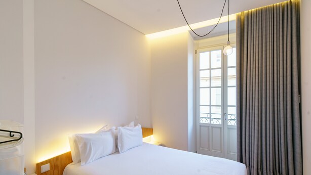 Habitación de hotel con una cama bien hecha, una mesita de noche con lámpara, una ventana con cortinas y vistas al exterior.