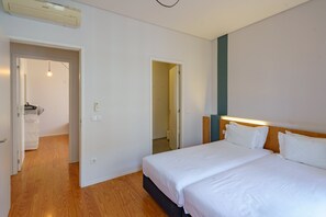 A hotel room with a wooden floor, a large bed with white bedding, a wooden headboard, and a wall-mounted air conditioning unit.