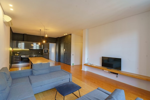 A modern living space with a kitchen, grey sofas, a wooden dining table, and a TV mounted on a wooden shelf.