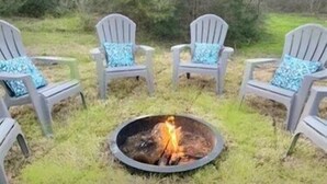 A circular fire pit surrounded by four Adirondack chairs with blue cushions, set in a grassy outdoor area. - NEAR BRAZOS COUNTY EXPO CENTER. ENJOY FRESH AIR AND STARRY NIGHTS. (Bryan)