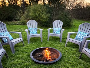 Three Adirondack chairs with blue floral cushions arranged around a circular fire pit on a grassy lawn. - Steps to Brazos County Expo 2 Acres + Fire Pit  Sleeps 9 (Bryan)