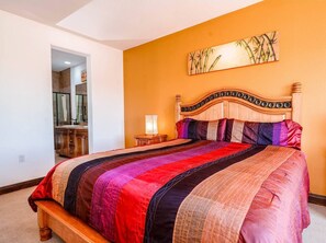 Ein Schlafzimmer mit einem hölzernen Bett, einem Nachttisch, einer Wandlampe und einem durch eine offene Tür einsehbaren Badezimmer.