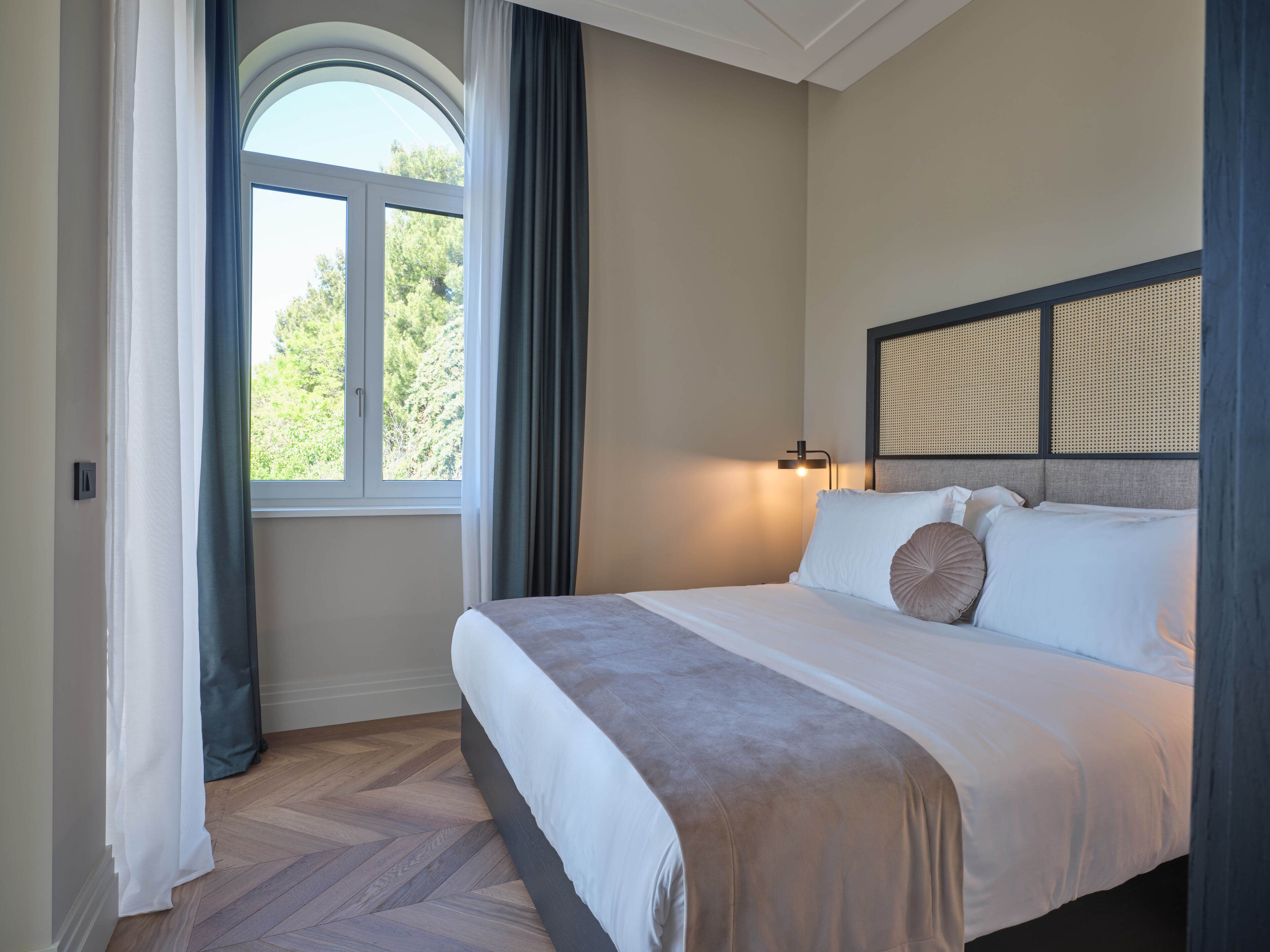 Ein ordentlich bezogenes Bett mit Kopfteil, ein Fenster mit Vorhängen und eine Wandleuchte.