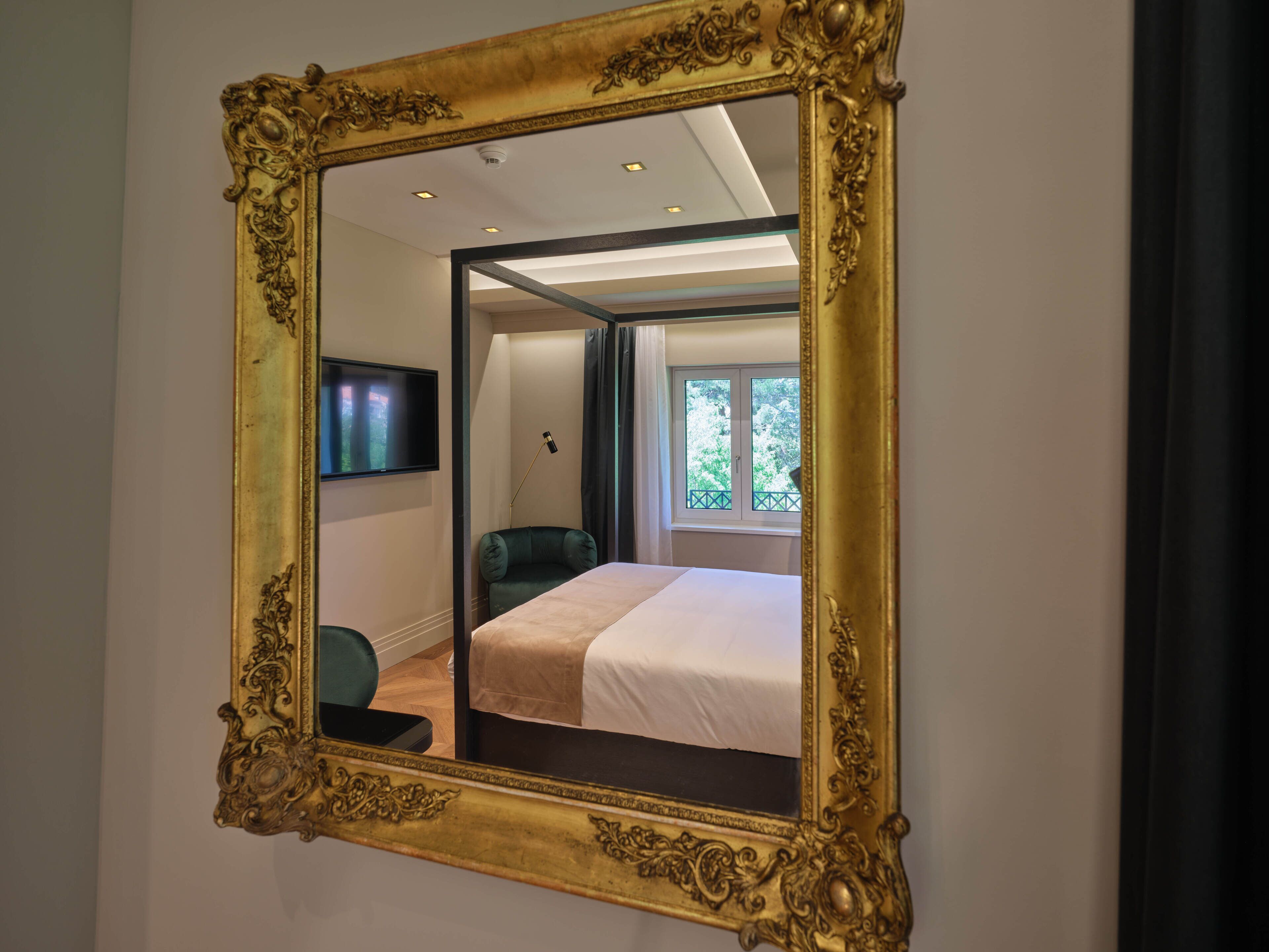 A bedroom with a large ornate mirror, a bed, a television, and a window with a view of trees.