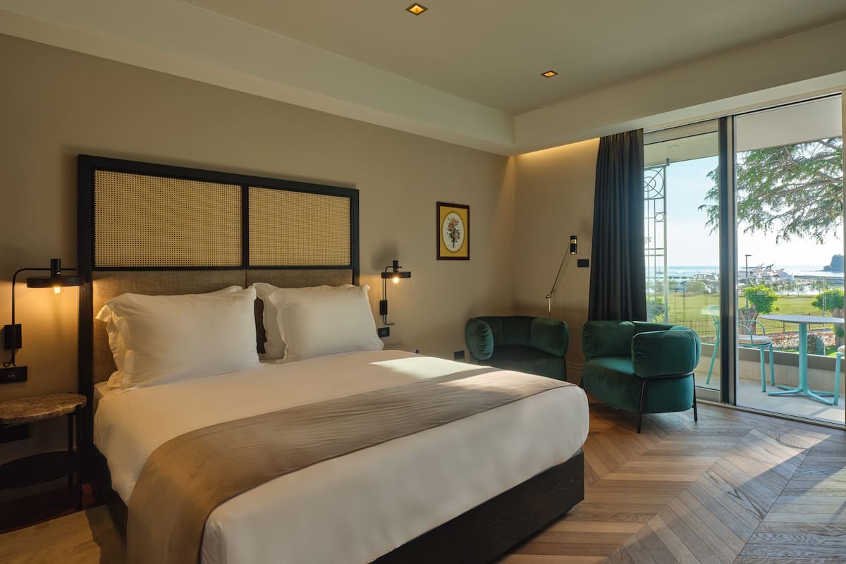 A modern hotel room with a large bed, a green armchair, and a balcony with a view of the city.