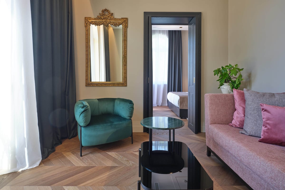 A modern living room with a teal armchair, a glass coffee table, a pink sofa, and a large mirror.