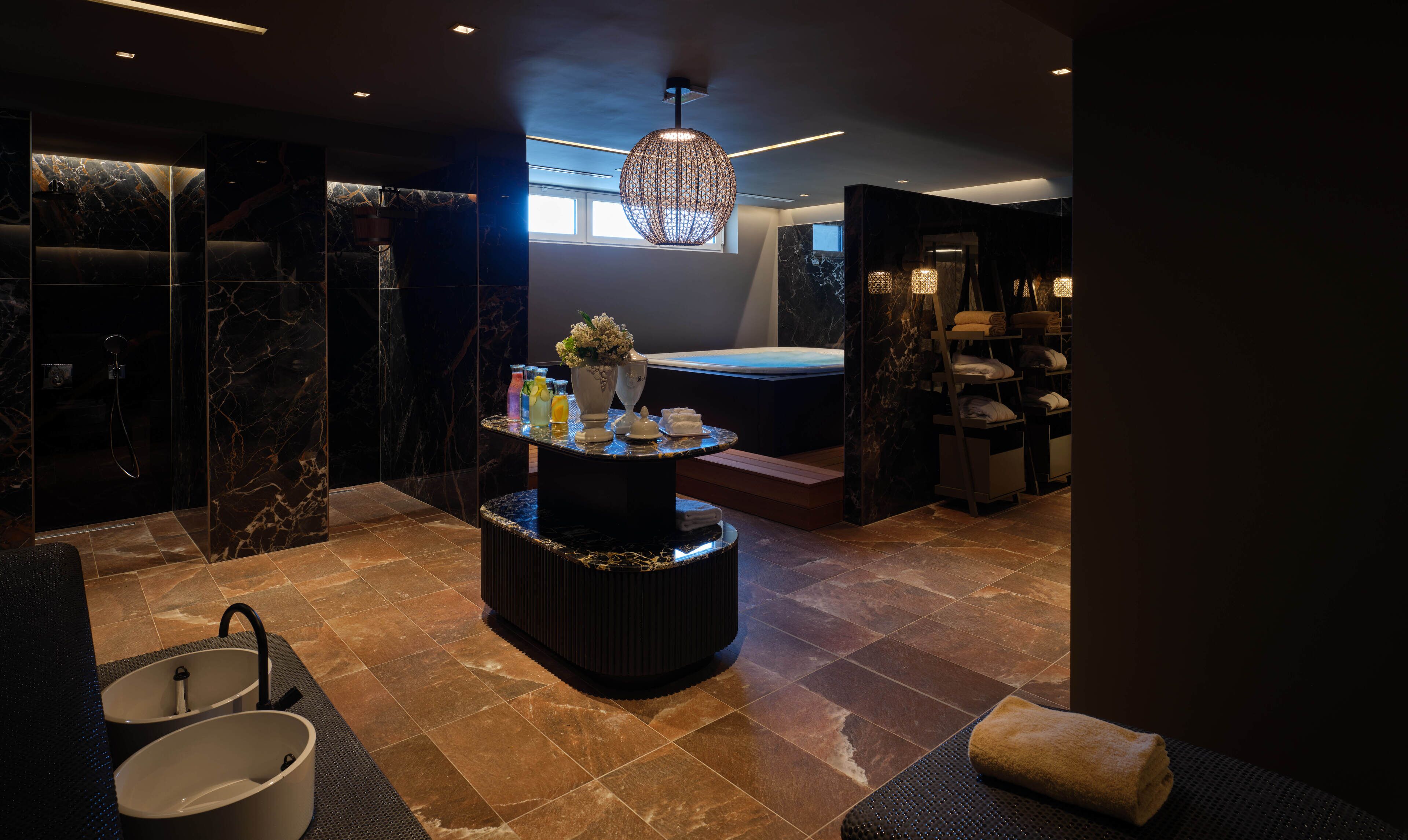A modern spa area with a marble floor, a central round table with amenities, a hot tub, and a large decorative light fixture.