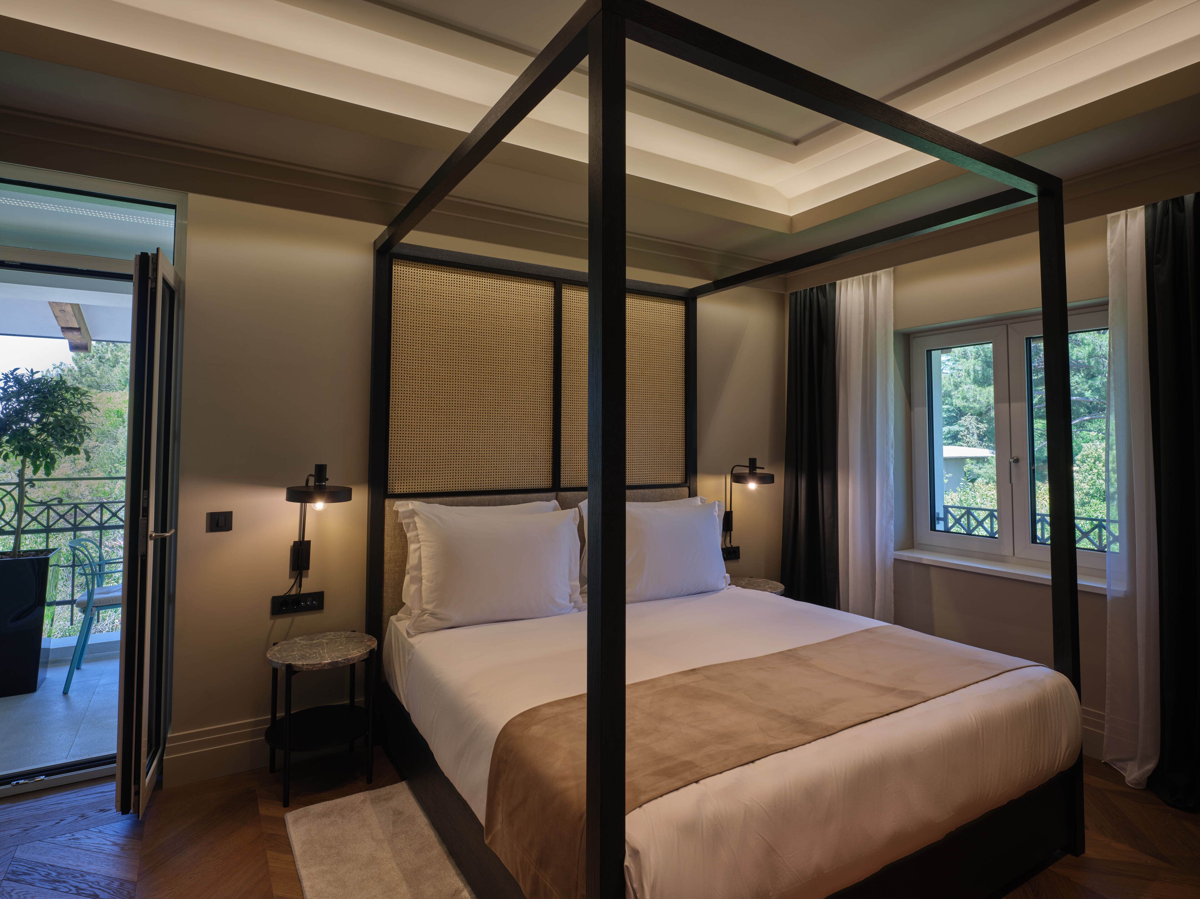 A four-poster bed with a canopy, a small bedside table, a nightstand, and a view of the outdoors through a window.