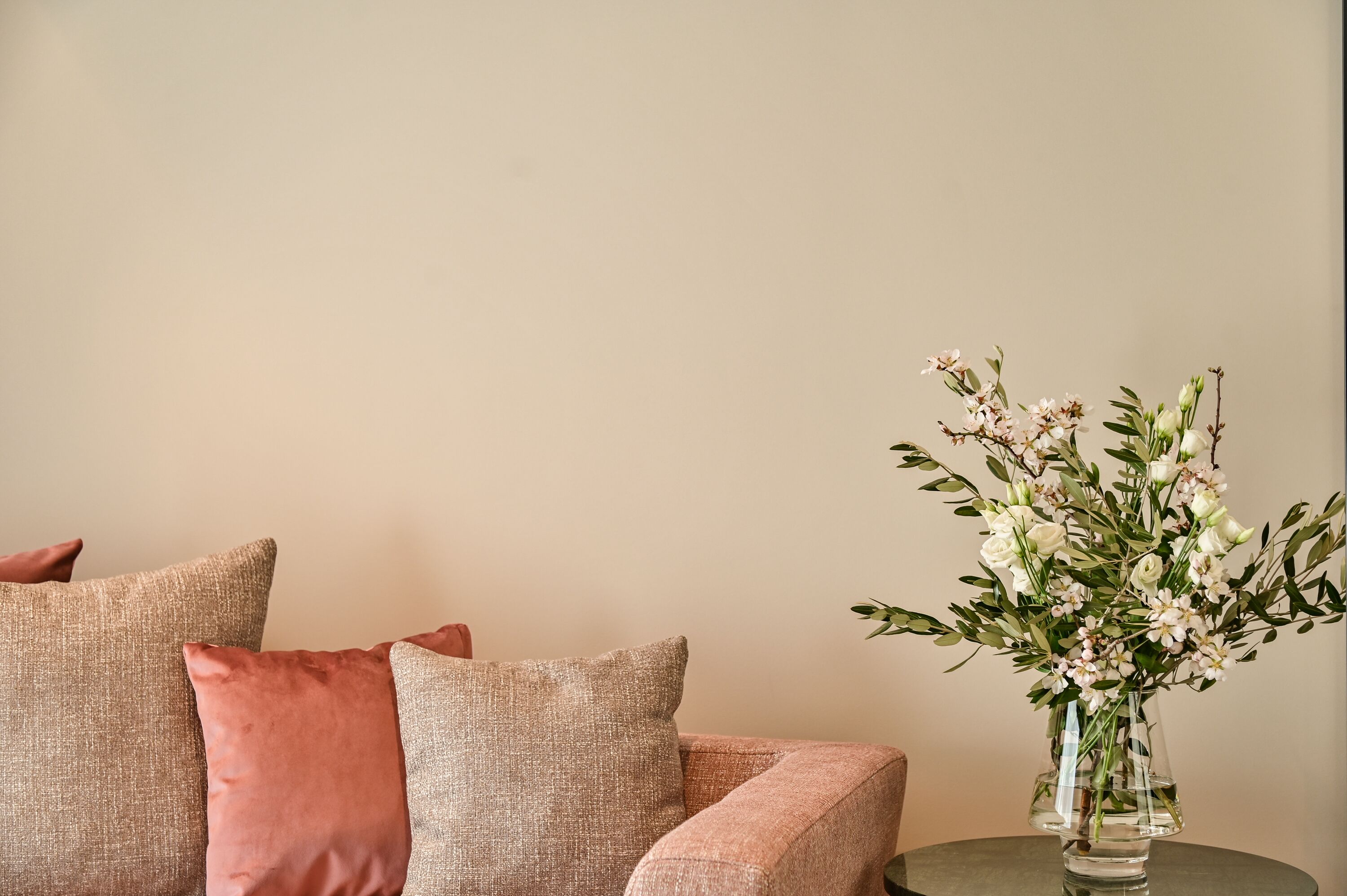 Um sofá bege com almofadas cor-de-rosa e um vaso transparente com flores brancas sobre uma mesa redonda.