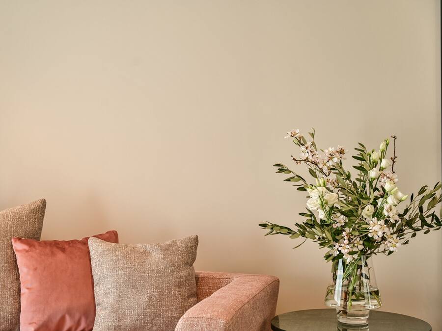 Um sofá bege com almofadas cor-de-rosa e um vaso transparente com flores brancas sobre uma mesa redonda.