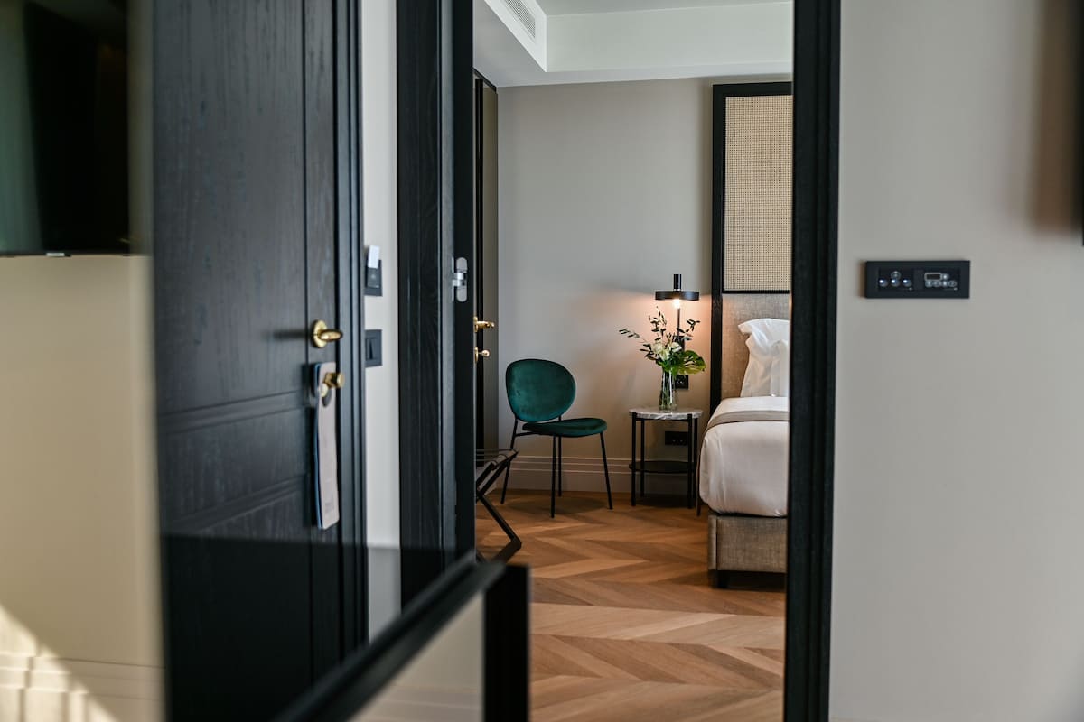 A hotel room with a wooden floor, a bed, a green chair, a small table with a vase of flowers, and a wall-mounted TV.