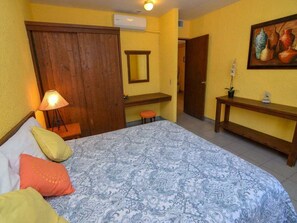 A bedroom with a bed, a nightstand, a lamp, a wooden wardrobe, a mirror, and a wall-mounted shelf.