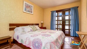 A bedroom with a bed, a nightstand, a desk, a window with curtains, and a painting on the wall. - La Hacienda Condo Six (San Felípe)