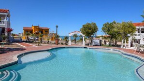 A swimming pool with a clear blue water feature, surrounded by a paved area with a white fence and a building with a red roof. - La Hacienda Condo Six (San Felípe)