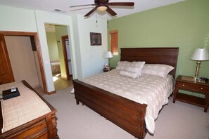 Um quarto com cama de madeira, ventoinha no teto, mesa de cabeceira com abajur e porta para outro cômodo.