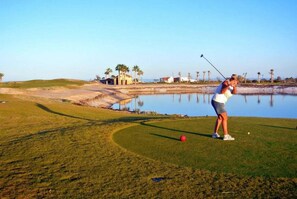 Un campo de golf con un cuerpo de agua, palmeras y un edificio al fondo.
