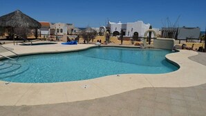 A swimming pool with a curved edge, a thatched-roof structure, and white buildings in the background. - Los Sahuaros House 34 (San Felipe)