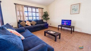 A living room with a sofa, a coffee table, a TV on a stand, a window with curtains, and a plant. - La Hacienda Condo 18 (San Felípe)