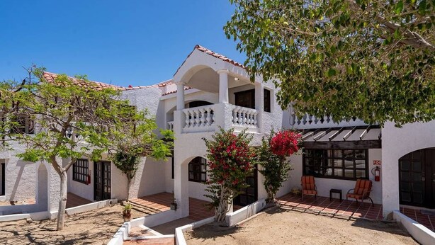 A white building with a tiled roof, a covered patio area with chairs, and a small garden with trees and flowers. - La Hacienda Condo 18 (San Felípe)