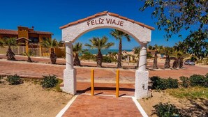 A white archway with the inscription "FELIZ VIAJE" leading to a beach area with palm trees and a clear blue sky. - La hacienda condo Four (San Felípe)