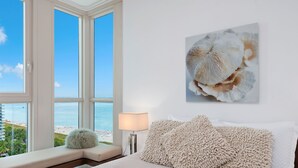 Een slaapkamer met een groot raam met uitzicht op een strand, een bed met zachte kussens en een lampje op een nachtkastje.