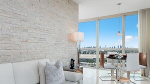 Een moderne woonkamer met een witte bakstenen muur, een witte bank met gekleurde kussens, een eettafel met stoelen, en een groot raam met uitzicht over de stad.