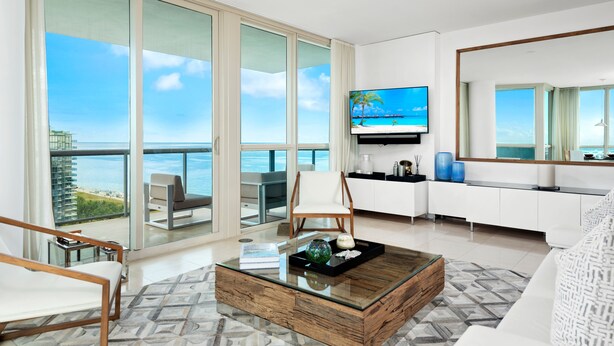 Een moderne woonkamer met een grote glazen deur die uitkomt op een balkon met uitzicht op zee. Er is een flatscreen televisie aan de muur bevestigd, een witte bank en een houten bijzettafeltje met decoratieve objecten.