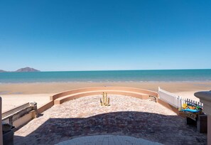 Vista de la playa con un paseo adoquinado que lleva al agua, una cerca blanca y una pequeña construcción con techo azul.