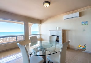 Uma área de refeições com mesa e cadeiras de vidro, lareira, ar condicionado e vista para o mar.