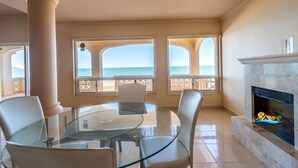 A room with a glass table and chairs, a fireplace, and a view of the sea through large windows.