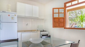 A modern kitchen with white cabinets, a glass-top dining table, and a window with a view of greenery. - IT16 Apt 2 Quartos a 150m da Praia (Camaçari)