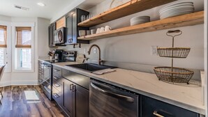 A modern kitchen with dark cabinetry, a stainless steel dishwasher, and wooden shelves holding plates and cups. - Ocean views includes Golf Cart - 611 Newhaven (Fripp Island)