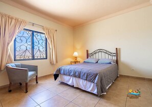 Un dormitorio con cama, una silla, una ventana con cortinas y una mesita de noche con lámpara.