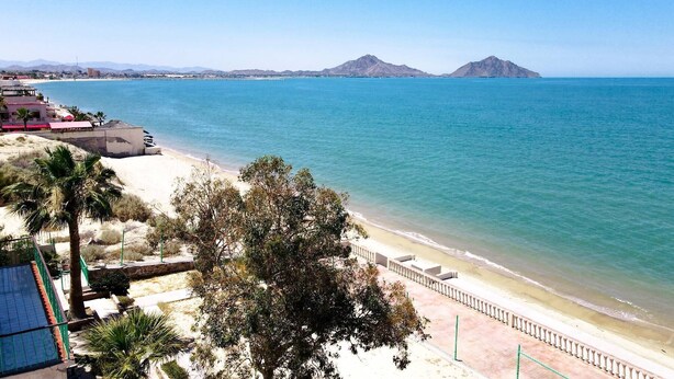A beach with a clear view of the ocean and distant mountains. There are buildings and a fence along the beach. - Jerry`s Beach Club Condo 2 (San Felipe)