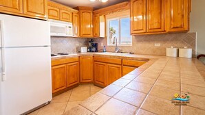 A kitchen with wooden cabinets, a white refrigerator, a microwave, and a window with a view of palm trees. - Jerry`s Beach Club Condo 2 (San Felipe)