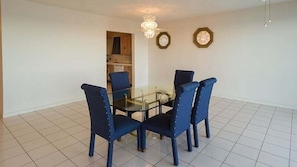 Een eetgedeelte met een glazen tafel en vier stoelen, een keuken op de achtergrond en een kroonluchter.