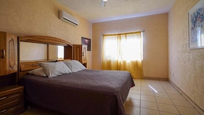 A bedroom with a bed, wooden headboard, curtains, and a wall-mounted air conditioner.