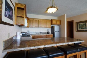 A kitchen with granite countertops, wooden cabinets, a built-in island with bar stools, a hanging light fixture, and a refrigerator.