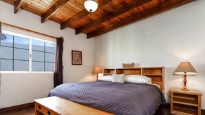 A bedroom with a wooden bed, a bench at the foot of the bed, a bedside table with a lamp, and a window with curtains. - Rick`s Private POOL House (San Felípe)