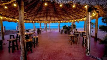 Un espace de restauration en plein air doté de toits de chaume et illuminé de guirlandes lumineuses, avec vue sur la plage.
