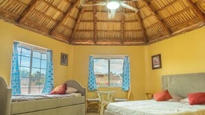 Una habitación con techo de paja, dos camas, una mesita y dos ventanas con cortinas.