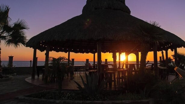 A thatched-roof pavilion with outdoor seating, overlooking a sunset over the ocean.