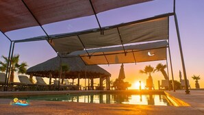 A pool area with lounge chairs and umbrellas, a thatched-roof structure, and a sunset in the background.