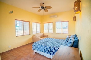 Quarto com cama, ventilador de teto e três janelas com persianas.