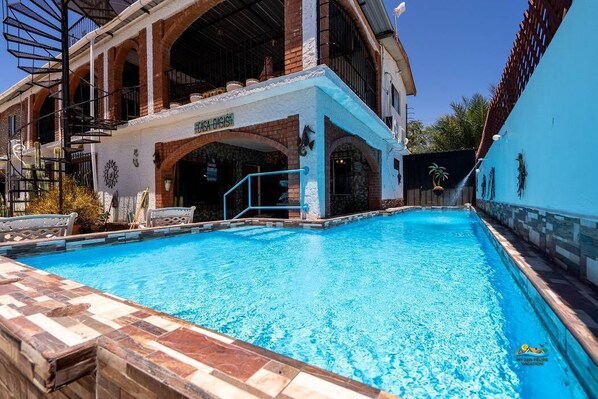 Une piscine avec une terrasse carrelée, un bâtiment en briques doté d’arcades et une enseigne sur laquelle on peut lire « CASA D’IS ».