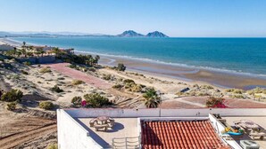Un hôtel situé sur la côte, doté d’une terrasse, offrant une vue sur la plage et ayant les montagnes en toile de fond.